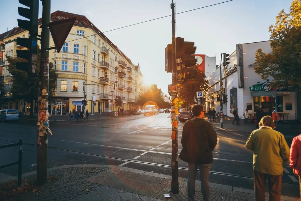 A sunlit street corner: two pedestrians wait to cross while a van drives towards them
